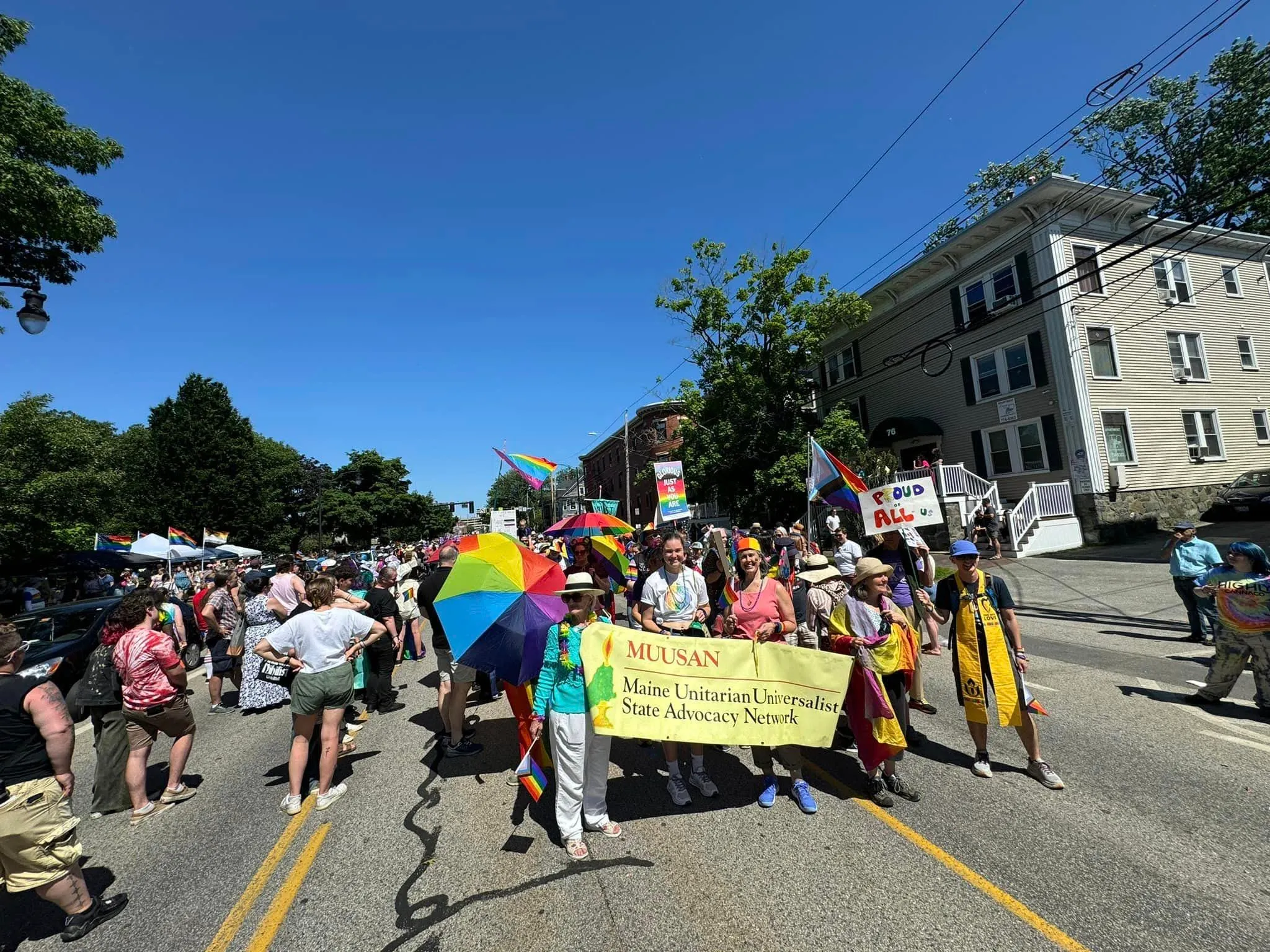 A2U2 members march at Pride behind the yellow MUUSAN banner, with a large rainbow umbrella under a blue sky